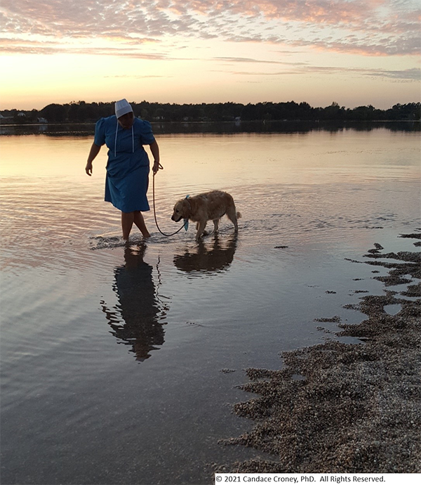 An Amish woman walks an older golden retriever on a leash at sunset in the water along the edge of a lake.